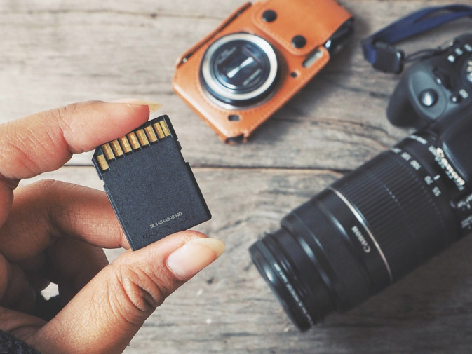 Hand holding a camera card above a table with 2 cameras on it to inspect for camera card data recovery.