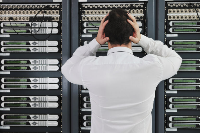 Man wearing a white shirt with hands held up to both sides of his head standing in front of RAID array equipment experiencing RAID Failure.